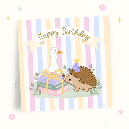 A square birthday card with pastel vertical stripes in pink, blue, yellow, and white, featuring an illustration of a white goose, a brown hedgehog wearing a purple party hat, and a stack of gift boxes tied with a yellow ribbon. A cream banner across the top reads 'Happy Birthday' in gold script.