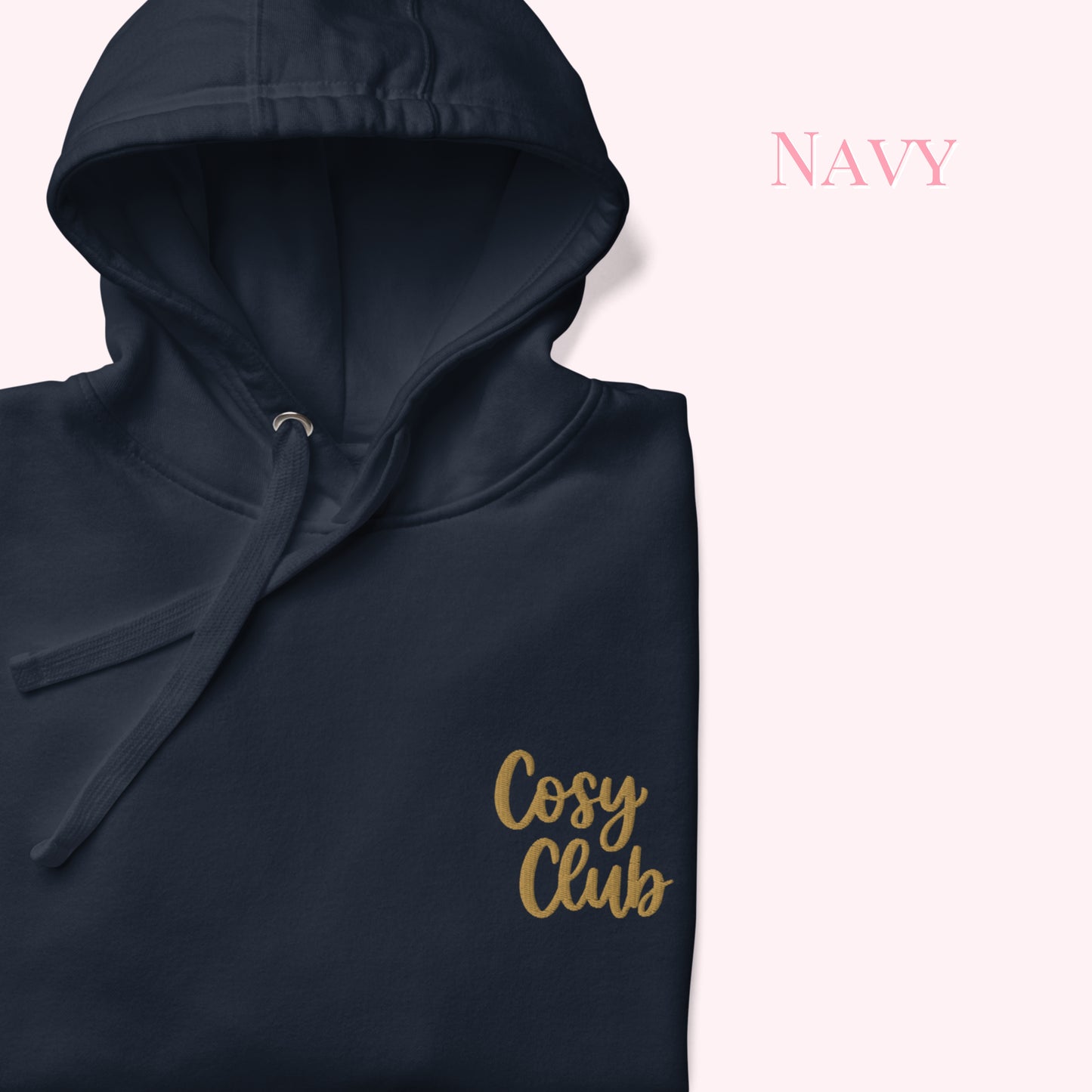 A folded navy hoodie with a gold 'Cosy Club' script logo on the front, displayed on a pale pink background with the word 'Navy' in pink text in the upper right.