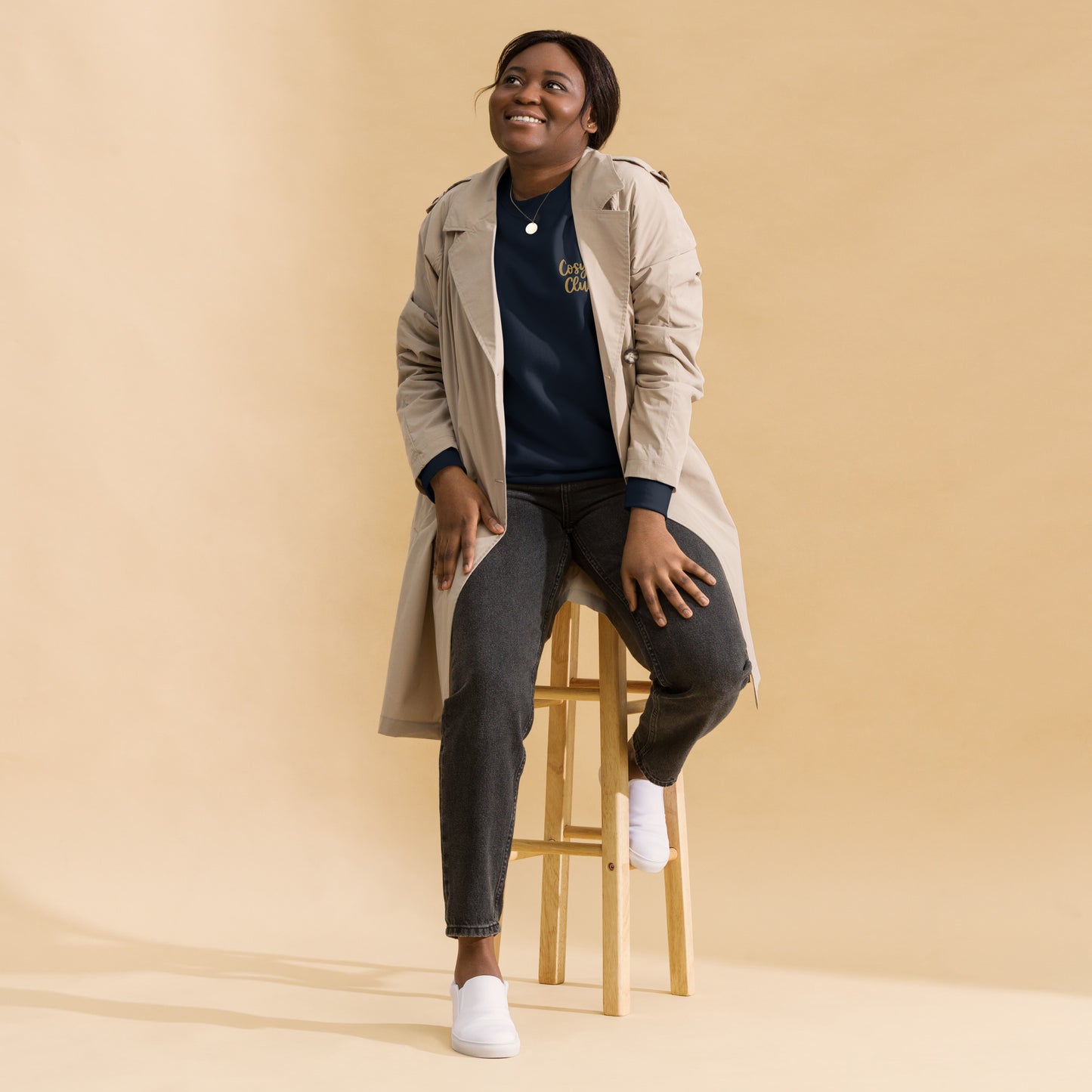 Woman sitting on a stool wearing a beige trench coat, black jumper with cosy club embroidered, and jeans against a beige background