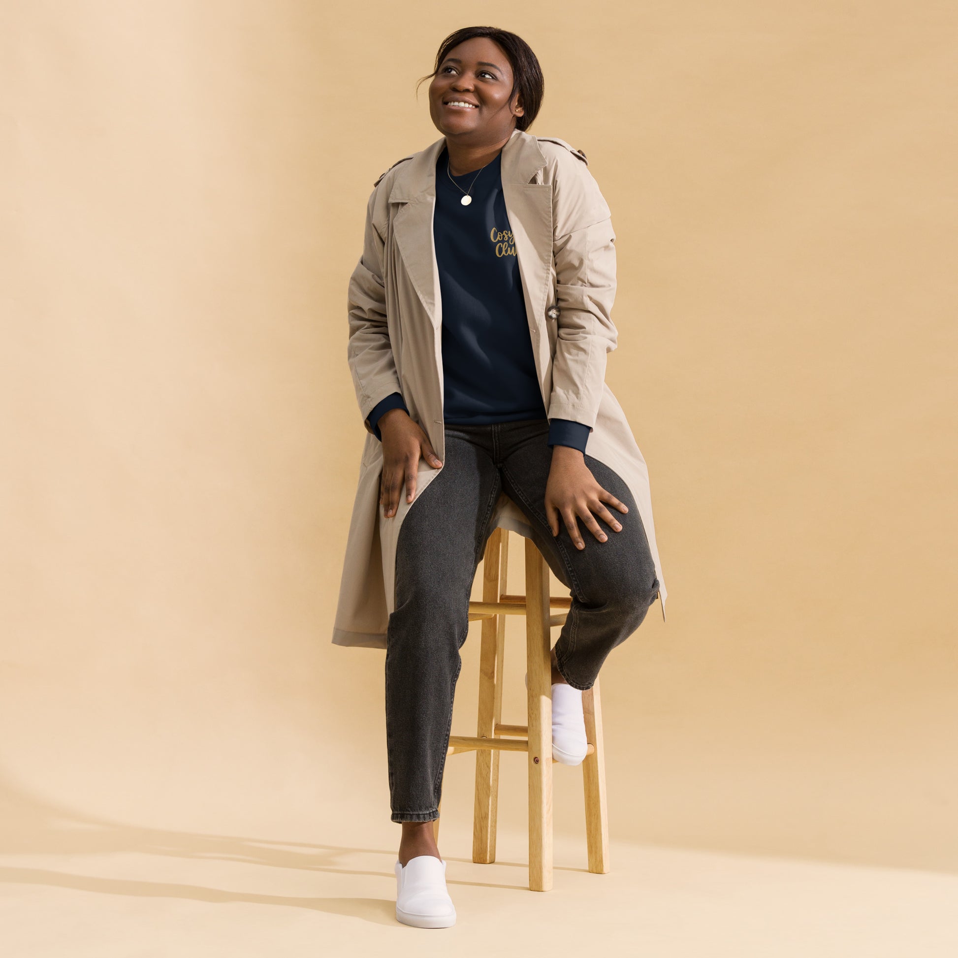 Woman sitting on a stool wearing a beige trench coat, black jumper with cosy club embroidered, and jeans against a beige background
